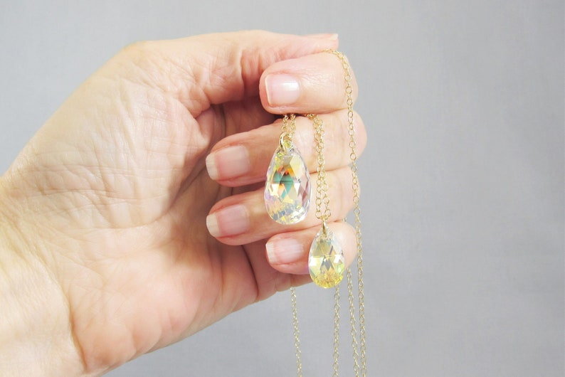 May include: Two gold necklaces with iridescent teardrop-shaped crystal pendants. The necklaces are layered and held in a hand.