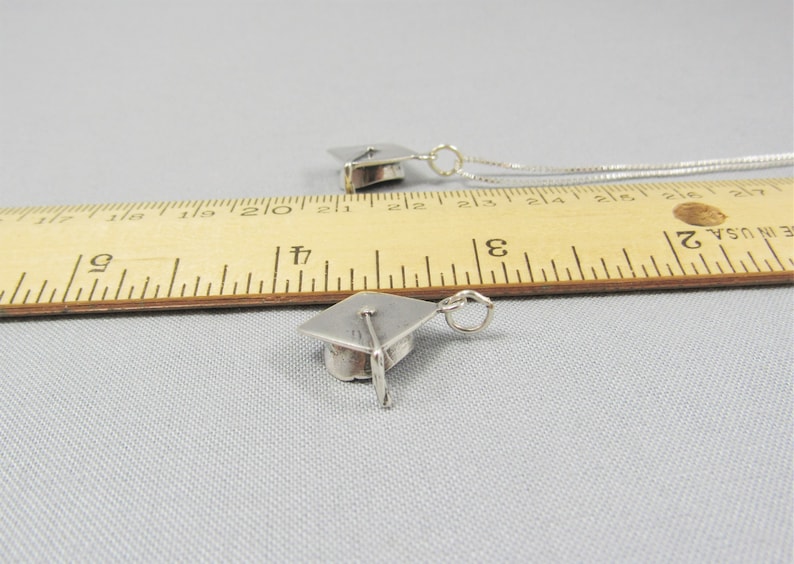 May include: Two silver graduation cap charms on a ruler. The charms are about 1 inch in size.