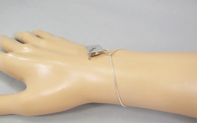 May include: A silver graduation cap charm on a silver chain bracelet. The bracelet is on a mannequin's wrist.