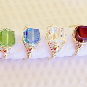 May include: Four wire-wrapped rings with different colored square-shaped crystals. The rings are silver and the crystals are green, blue, clear, and red.