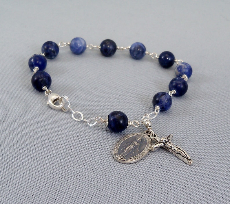 May include: A blue gemstone bracelet with a silver clasp and a silver charm featuring the Virgin Mary and a crucifix.