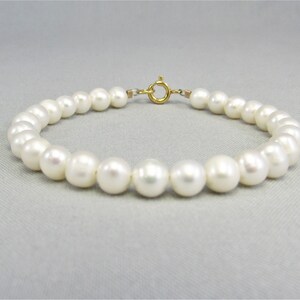 May include: A delicate bracelet featuring a strand of lustrous, round white pearls. The pearls are strung together with a gold-colored clasp. The bracelet is displayed on a light gray surface, showcasing its elegant design.