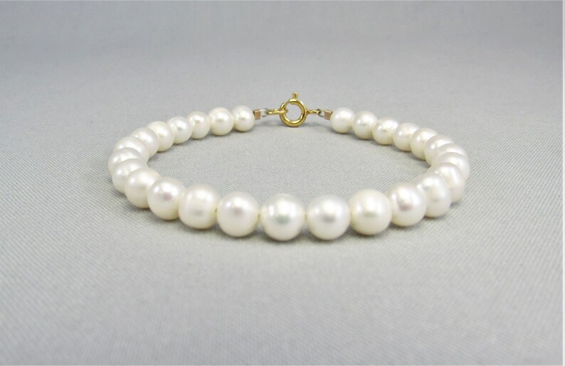 May include: A delicate bracelet featuring a strand of lustrous, round white pearls. The pearls are strung together with a gold-colored clasp. The bracelet is displayed on a light gray surface, showcasing its elegant design.