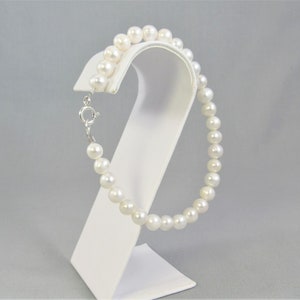 May include: A delicate pearl bracelet with a silver clasp, displayed on a white stand. The bracelet features a strand of round, white pearls. The clasp is silver and secures the bracelet.