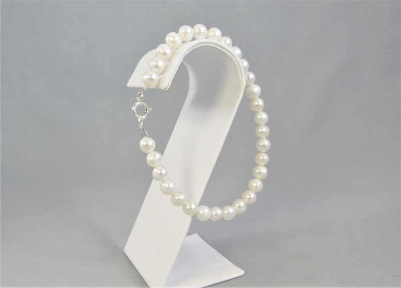 May include: A delicate pearl bracelet with a silver clasp, displayed on a white stand. The bracelet features a strand of round, white pearls. The clasp is silver and secures the bracelet.