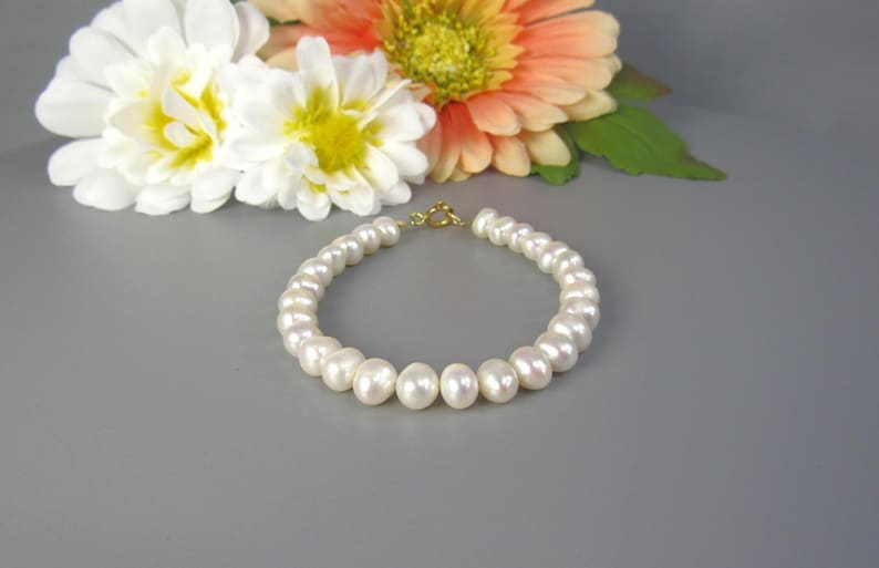 May include: A white pearl bracelet with a gold clasp. The bracelet is composed of round, lustrous pearls. White and orange flowers are in the background.