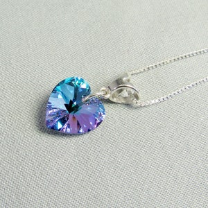 May include: A heart-shaped pendant necklace with a shimmering, iridescent crystal in shades of blue and purple. The pendant is suspended from a delicate silver chain. The jewelry is displayed on a light gray surface.
