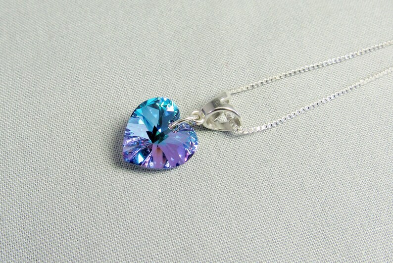 May include: A heart-shaped pendant necklace with a shimmering, iridescent crystal in shades of blue and purple. The pendant is suspended from a delicate silver chain. The jewelry is displayed on a light gray surface.