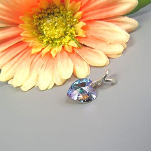 May include: A heart-shaped pendant with a shimmering, iridescent finish, suspended from a silver-toned bail. The pendant is set against a soft gray background, next to a large, orange and yellow gerbera daisy with green leaves.