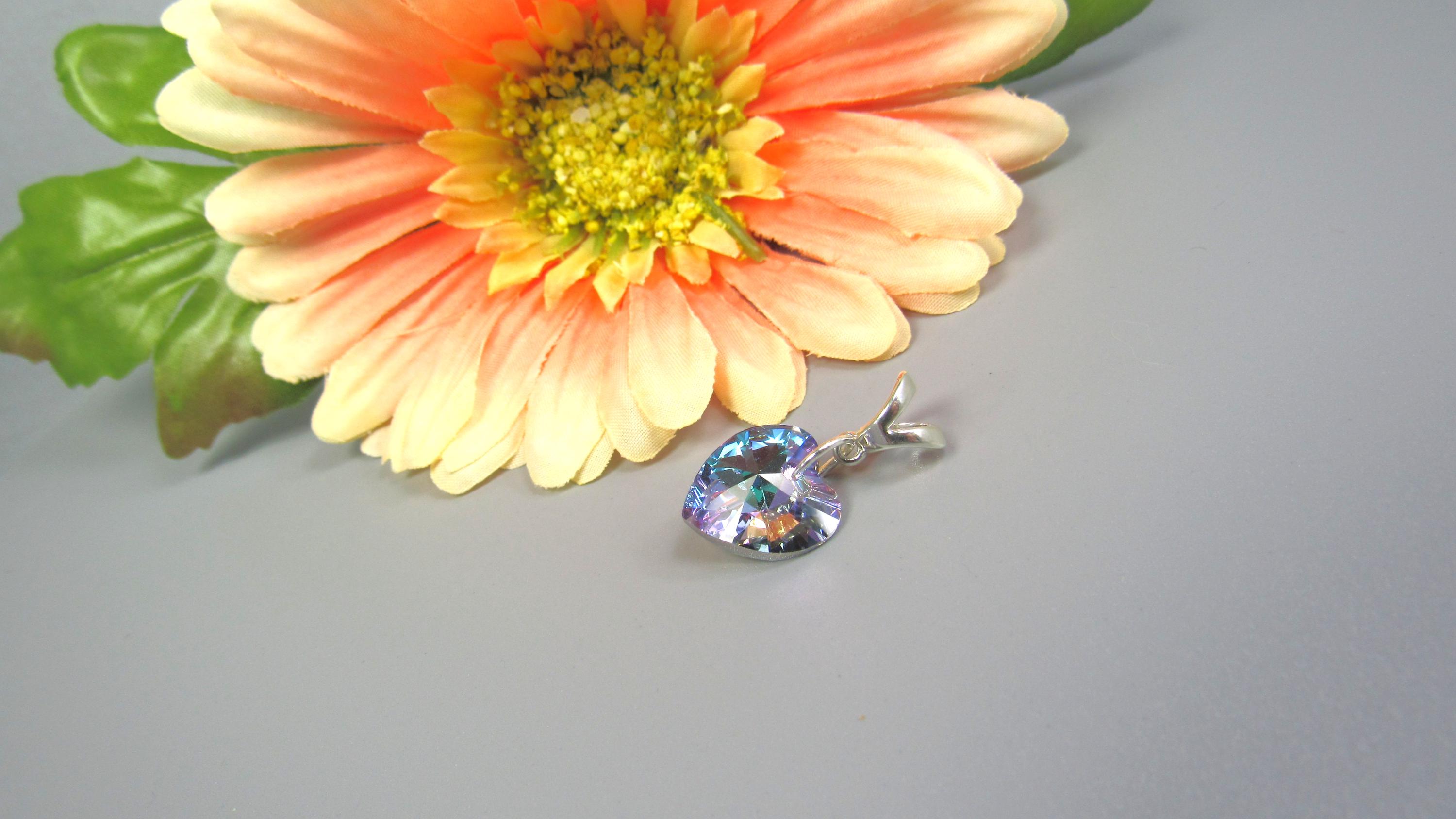 May include: A heart-shaped pendant with a shimmering, iridescent finish, suspended from a silver-toned bail. The pendant is set against a soft gray background, next to a large, orange and yellow gerbera daisy with green leaves.