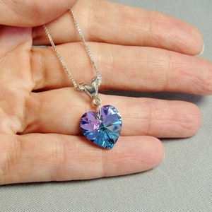 May include: A silver necklace with a heart-shaped pendant. The pendant is a faceted crystal with shades of blue and purple. The necklace is held in a hand against a gray background.