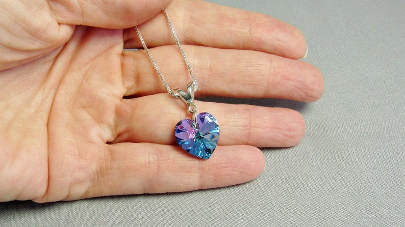 May include: A silver necklace with a heart-shaped pendant. The pendant is a faceted crystal with shades of blue and purple. The necklace is held in a hand against a gray background.