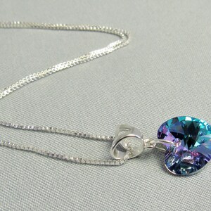 May include: A silver necklace with a heart-shaped pendant. The pendant is a vibrant mix of blue, green, and purple hues. The chain is a delicate, silver-toned design. The necklace is displayed on a light gray background.