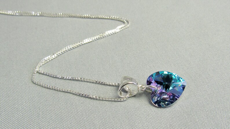 May include: A silver necklace with a heart-shaped pendant. The pendant is a vibrant mix of blue, green, and purple hues. The chain is a delicate, silver-toned design. The necklace is displayed on a light gray background.