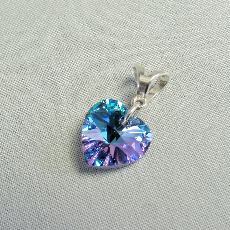 May include: A heart-shaped pendant with a faceted crystal in shades of blue and purple. The pendant is suspended from a silver-colored bail. The crystal reflects light, creating a sparkling effect. The background is a light gray fabric.