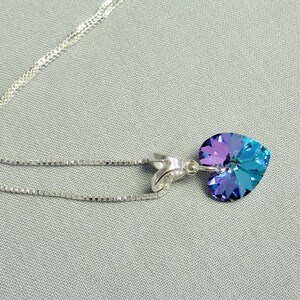 May include: A silver necklace with a heart-shaped pendant. The pendant is a gradient of purple and teal. The chain is a delicate, silver box chain. The necklace is displayed on a light gray background.