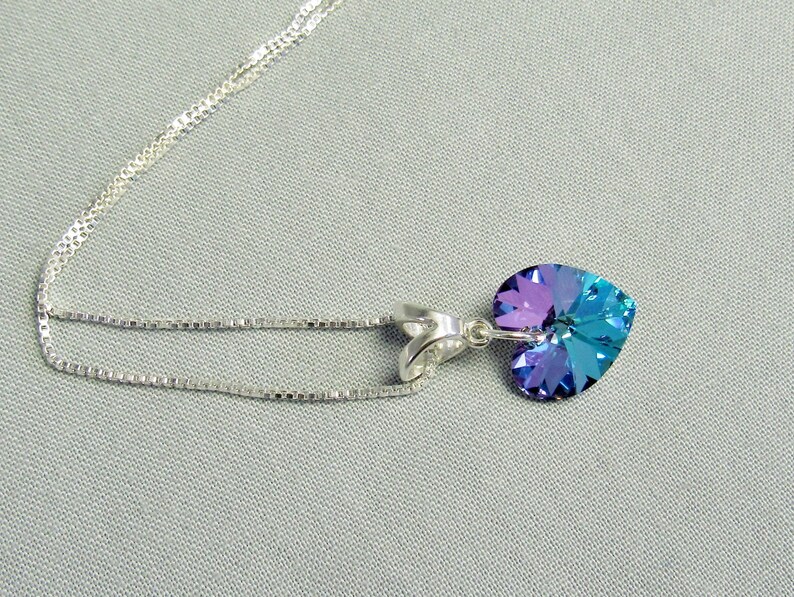 May include: A silver necklace with a heart-shaped pendant. The pendant is a gradient of purple and teal. The chain is a delicate, silver box chain. The necklace is displayed on a light gray background.