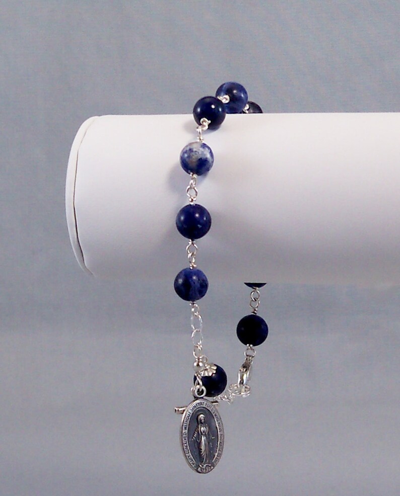 May include: A silver chain bracelet with blue and white gemstone beads and a silver charm of the Virgin Mary.