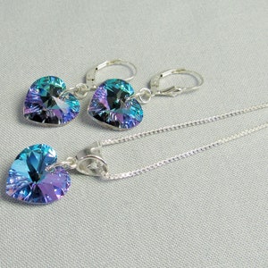 May include: A jewelry set featuring a heart-shaped pendant necklace and matching earrings. The pendant and earrings have iridescent blue and purple crystals. The necklace and earrings are made of silver-colored metal.