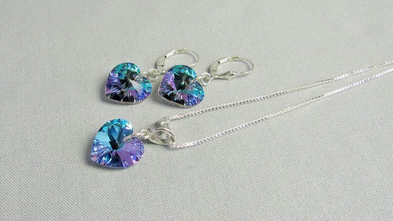 May include: A jewelry set featuring a heart-shaped pendant necklace and matching earrings. The pendant and earrings have iridescent blue and purple crystals. The necklace and earrings are made of silver-colored metal.
