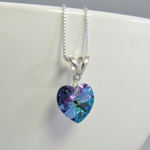May include: A heart-shaped pendant necklace with a silver chain. The pendant is a large, iridescent crystal with shades of blue, purple, and green. The necklace is displayed against a white background.