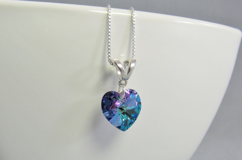 May include: A heart-shaped pendant necklace with a silver chain. The pendant is a large, iridescent crystal with shades of blue, purple, and green. The necklace is displayed against a white background.