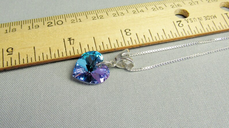 May include: A heart-shaped pendant necklace with a silver chain. The pendant is a gradient of blue and purple. A wooden ruler with inch markings is in the background.