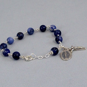 May include: A silver chain bracelet with blue gemstone beads and a silver charm with a religious figure and a cross.