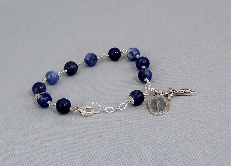 May include: A silver chain bracelet with blue gemstone beads and a silver charm with a religious figure and a cross.