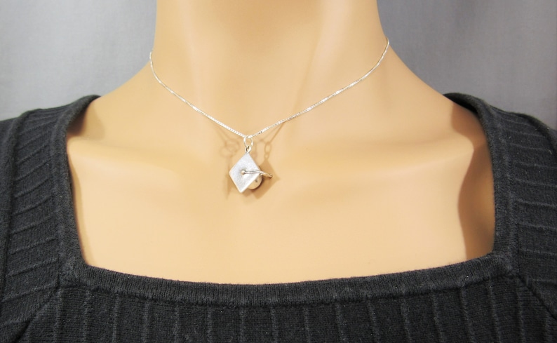 May include: A silver chain necklace with a small silver pendant shaped like a graduation cap. The pendant has a small pearl in the center.