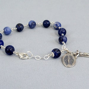 May include: A blue gemstone bracelet with a silver chain and a silver charm featuring the Virgin Mary and a cross.