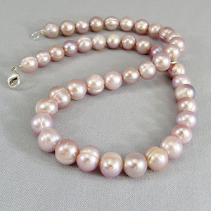 May include: A necklace composed of round, light pink pearls. The pearls are strung together with a silver clasp. The necklace is displayed on a gray surface. The pearls have a lustrous sheen.