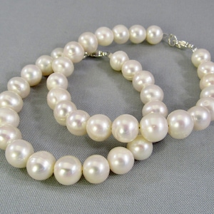 May include: A strand of lustrous white pearls arranged in a circular shape, featuring smooth, spherical beads with a reflective surface and a silver-toned metal clasp mechanism visible at the top where the two ends of the strand meet.