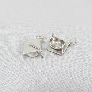 May include: Two silver graduation cap charms. One charm is a flat, square shape with a raised graduation cap design. The other charm is a three-dimensional graduation cap.