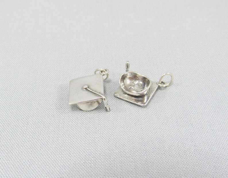 May include: Two silver graduation cap charms. One charm is a flat, square shape with a raised graduation cap design. The other charm is a three-dimensional graduation cap.