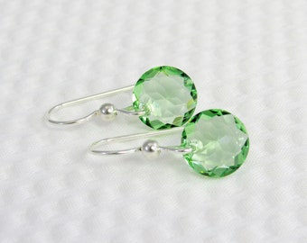 Peut inclure: Une paire de boucles d'oreilles avec des pierres précieuses rondes et facettées, de couleur vert clair. Les pierres sont suspendues à des crochets d'oreilles argentés incurvés avec de petites perles argentées. Sur fond blanc texturé.