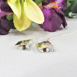 May include: A silver necklace with two graduation cap charms. The necklace is on a white surface with purple and yellow flowers in the background.