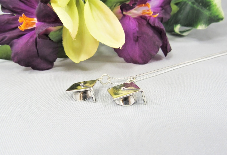 May include: A silver necklace with two graduation cap charms. The necklace is on a white surface with purple and yellow flowers in the background.