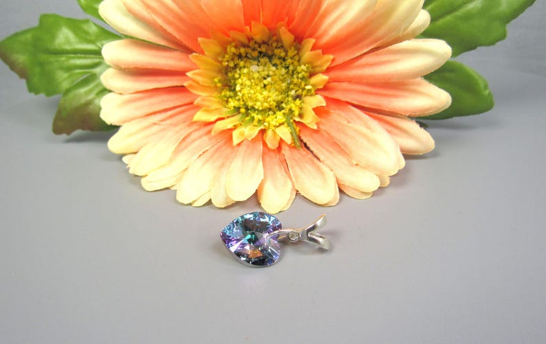 May include: A heart-shaped pendant with iridescent colors and a silver-tone bail. The pendant is set against a gray background, with a large orange and yellow flower in the background.