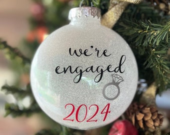 May include: A white glitter Christmas ornament with the text "we're engaged 2024" and a black and white ring with a diamond.