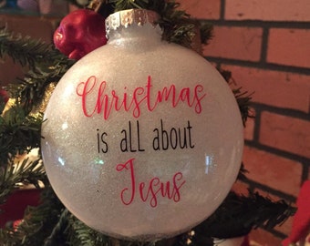 May include: White glitter Christmas ornament with the text "Christmas is all about Jesus" in red lettering.