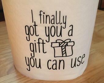 May include: A white roll of toilet paper with the text "I finally got you a gift you can use" printed on it. A small drawing of a gift box is included in the text.