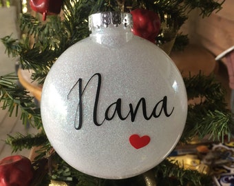 May include: White glitter Christmas ornament with the word "Nana" in black script and a red heart.