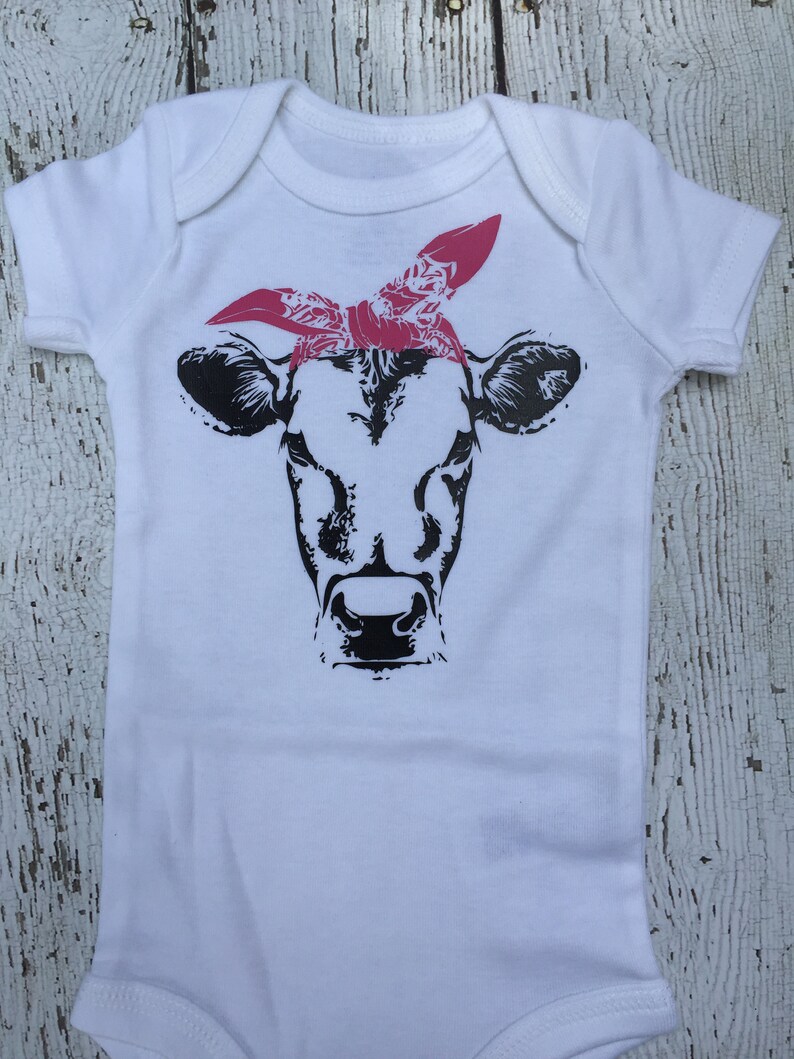 May include: White baby onesie with a black and white cow wearing a pink bandana.