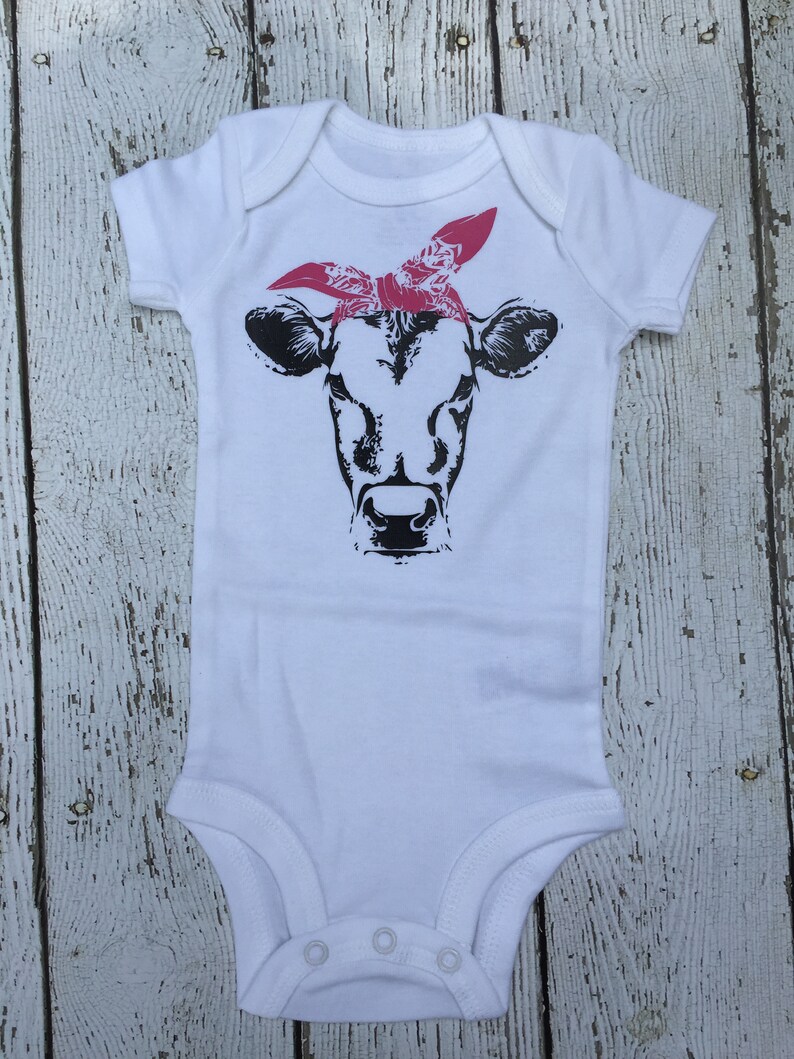 May include: White baby onesie with a black and white illustration of a cow wearing a pink bandana.