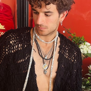 May include: A black open-front cardigan with a textured, fringed knit design. The garment is worn with a long, shimmering silver sequined scarf draped around the neck and a layered necklace set featuring black beads, silver chains, and metal pendants.