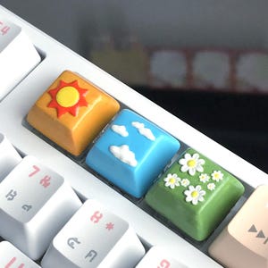 以下が含まれることがあります： 白いキーボードに、カスタムキーキャップが4つ付いています。キーキャップは、太陽、雲、デイジー、再生ボタンで飾られています。キーキャップは、柔らかくカラフルな素材で作られています。