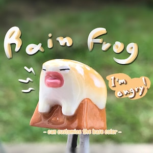 May include: A small ceramic frog figurine with a yellow and orange glaze. The frog has a pink mouth and is painted with black eyes. The frog is sitting on a brown base and has the words "Rain Frog" and "I'm angry!" written in white on the background.