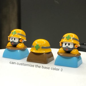 May include: Three yellow and black robot figurines wearing yellow hard hats with green crosses. The robots are sitting on blue and brown bases. "can customize the base color." is written below the robots.