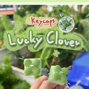 May include: Two square light green mechanical keyboard keycaps featuring a raised four-leaf clover design with small white eyes and a tiny red mouth. The text Keycaps Lucky Clover is displayed at the top of the image in a decorative font.